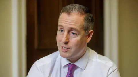 PA Media Paul Givan. He has shirt grey-ish hair and blue eyes, wearing a white shirt and purple paisley tie. A brown wooden door and yellow wall is blurred in the background.