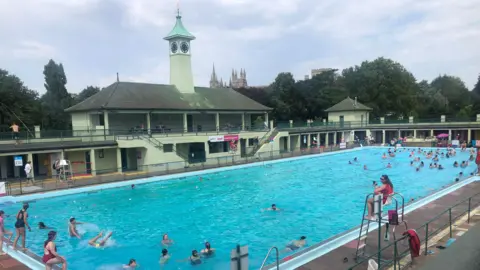 Swimmers dip in the blue water of Peterborough's outdoor swimming pool, while a lifeguard watches on. In the background, the art deco clock turret of the lido's entrance can be seen. 