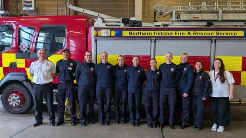 A male firefighter wearing a white shirt and black trousers alongside 10 female fire fighters. A fire engine is behind them.