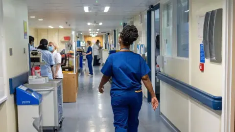 PA Media A nurse wearing blue scrubs walks down a hospital corridor, she has her hair up in a bun.