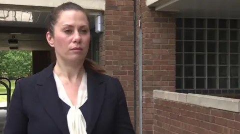 Det Ch Insp Becky Woodsford, a woman wearing a smart navy jacket with a white blouse and has dark hair in a ponytail. She is standing in a housing estate