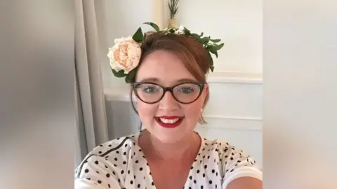 Handout A photo of a smiling woman who is wearing a white t-shirt with black polka dots, black framed glasses, bright red lipstick and a headband with flowers on. She is photographing herself from the shoulder up.