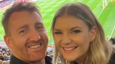 Siobhan Gorman A selfie of Siobhan and her partner who are in a rugby stadium. Both are smiling at the camera from the seats with the pitch behind them. She has long wavy blonde hair and is smiling. She is in an office. He has short ginger hair and a cropped beard.