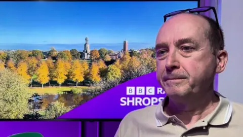 A man with short brown hair, a pale shirt and glasses perched on top of his head in front of a purple illuminated background and am image of tall buildings in between trees