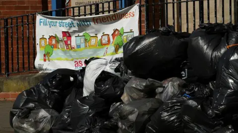 Reuters Bin bags are stacked up in a heap next to a community sign that reads "fill your street with friendliness". The sign has pictures of houses and trees and is brightly coloured.