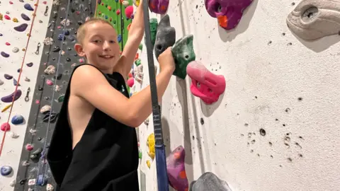 A boy with short blonde hair and wearing a black, sleeveless top, smiles as he holds onto handholds while scaling an indoor climbing wall.