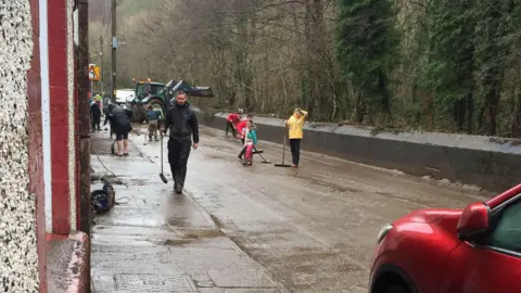 Paul Thomas Residents in Ynysybwl cleaning up after the flood, all have brushes in their hands brushing the mud out of the road. There is a tractor in the back of the image.