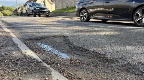 Pothole in a surburban road with two black cars parked by the kerb