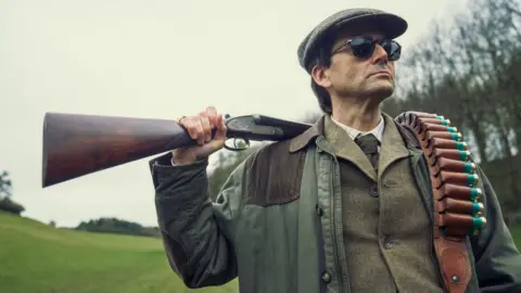 Disney+ David Tennant as Lord Tony Baddingham in Rivals, wearing sunglasses, a flat cap, and a tweed jacket as he holds a hunting rifle over his shoulder, resting ammunition on the other