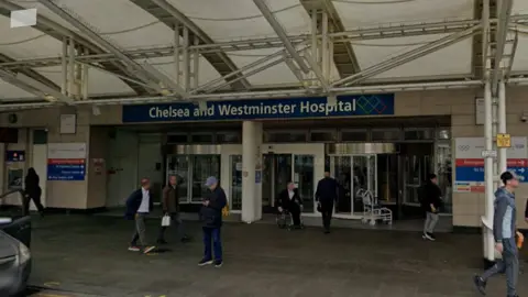 Google The entrance to Chelsea and Westminster Hospital, which opens onto a row of shops under a white canopy and has a blue sign. Members of the public walking past.