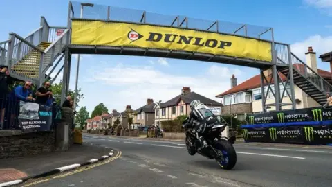 Michael Dunlop racing under the metal bridge with his front wheel off the ground. There is a large yellow Dunlop Tyres banner along the side of the structure overhead.