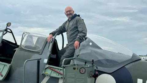 Brain Tumour Research The image shows a man sitting in what looks like a plane, smiling at the camera.