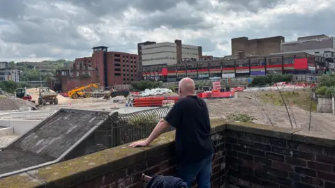 Simon Thake/BBC A bald man in a black t-shirt stands with his back to us looking out on a large excavation site with several workers and diggers