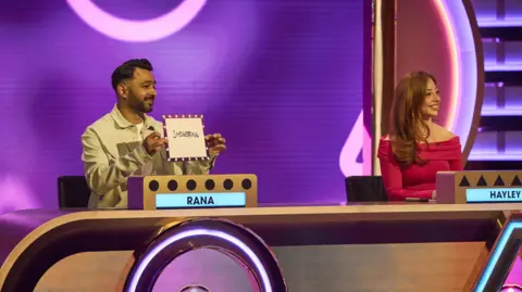 Fremantle/BBC Two contestants sit behind a brightly lit game show desk on a colourful studio set, each looking to the right while a man holds a card with handwriting. The set features purple and pink lighting, geometric patterns and name panels on the desk, giving the scene a lively television game show atmosphere.