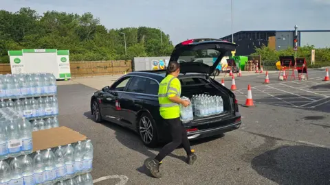 Michael Keohan/BBC A woman wearing a yellow hi-visibility jacket is carrying a pallet of water bottles and placing it in the back of a black car. Orange cones placed across the car park.