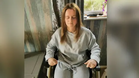 Melonie Bray Ms Bray sat in a wheelchair, she has her hands clenched and eyes and mouth closed. She is wearing a grey lounge-wear set. 