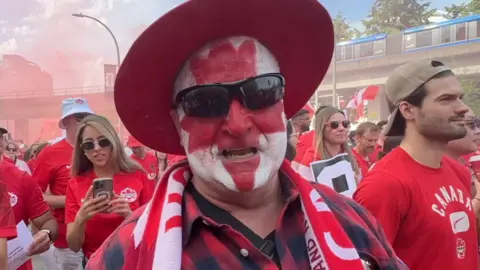 A man with his face painted to depict the Canadian flag leaf, wears a red hat and sunglesses.