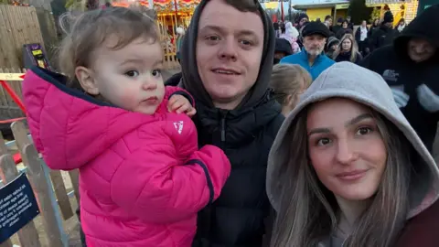 Alaw Llyfni Selfie of Alaw, Gwion and Nanw at a fun fair. Alaw, on the right, wears a grey hoodie. Gwion, stood next to Alaw wears a black hooded jacket and holds Nanw who is wearing a bright pink coat. 