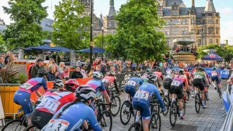 Many professional cyclists in lycra cycle down a temporary track which has been set up in a city centre. The town hall is visible in the background. Crowds of people line the route.