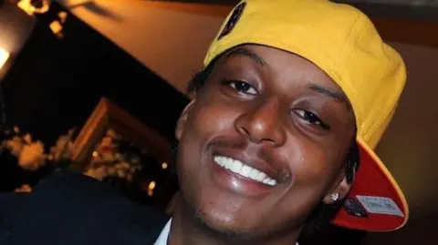 Wearing a yellow snapback hat, a close up photo shows Eddie Kinuthia smiling at the camera in a dark room.