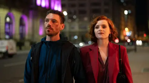 BBC/Left Bank Pictures/James Stack Filming of This City is Ours with Michael Kavanagh, played by James Nelson-Joyce, with short brown hair and beard, and Diana Williams, played by Hannah Onslow, with shoulder length red hair wearing a red blazer and burgundy top. There are walking in the street, in the evening, holding hands.