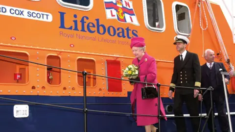 RNLI/ Simon Culliford Her Majesty Queen Elizabeth II going aboard Richard Cox Scott accompanied by Falmouth’s harbour master captain Mark Sansom and RNLI chairman Peter Nicholson on 1 May 2002. They are walking along the lifeboat which is orange and blue.