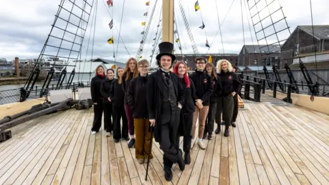 An actor as Isambard Kingdom Brunel, wearing a top hat and black dress attire with a waistcoat, and a cain. He is standing in front of around 10 of volunteers wearing black SS Great Britain jumpers, all standing in a v formation. They are on the deck of the SS Great Britain, with the rigging and water in the background. 