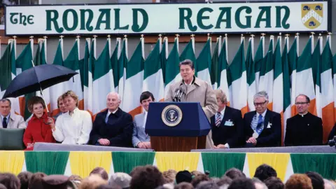 Bryn Colton/Hulton Archive/Getty Images The late US President Ronald Reagan addressing a crowd in Ballyporeen, County Tipperary in June 1984. He has short black hair and is wearing a beige raincoat. A large pub sign saying The Ronald Reagan is behind him, along with a line of Irish flags. Several people are on a stage behind him including his wife Nancy who is being sheltered by a woman holding an umbrella.