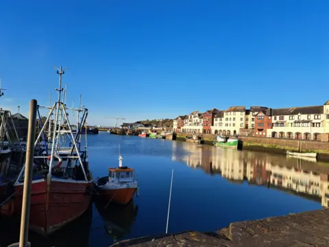 Maryport Marra Fishing boats at Maryport marina. Flats on one side of the dock cast reflections on to the water. The sky is blue without any clouds. 