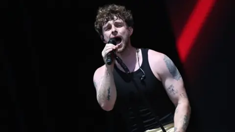 BBC Tom Grennan performing on stage in a black tank top and cream trousers.
