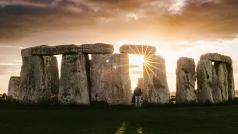  Man stands at Stonehenge with sunset and sunrays wonder - stock photo