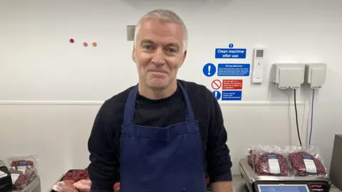 Leanne Rinne/BBC A man with white hair standing in front of a kitchen wall smiling. He is wearing a blue apron and a black jumper. You can see packets of venison meat on a table behind him. 