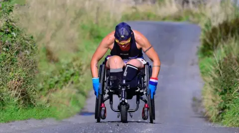 Lexi on her End2End challenge. She's in her wheelchair wearing a hat and blue gloves on a country lane going uphill. Green hedges either side