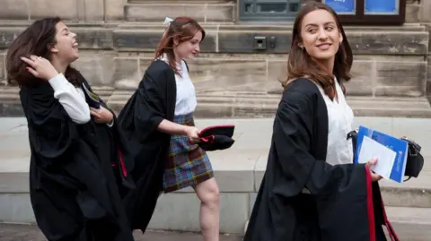 Getty Images Three young women wearing black gowns and formal dress walk outside a stone building. 