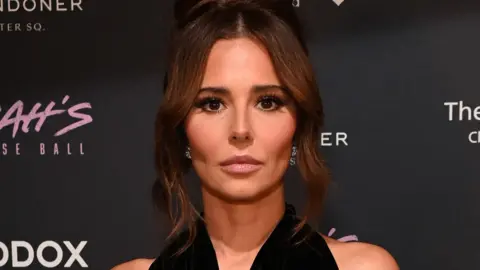 Getty Images Cheryl Tweedy: the head and shoulders of a woman in front of a black background which is marked with advertising slogans. Her face is framed by brown hair, which is touching her the bottom of her neck. She has brown eyes and is wearing silver earrings and what appears to be a black off-the shoulder dress.  