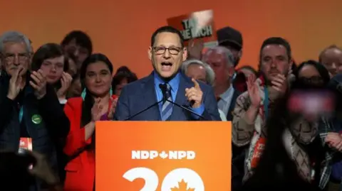 An image showing Avi Lewis speaking at an orange podium with the NDP logo. He is on a stage surrounded by supporters who are cheering him on.
