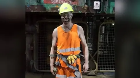 Supplied Shane Harrison wears a yellow hard had, clear glasses and orange hi-vis clothing. He also has a utility belt with keys and gloves attached. His face is dirty from being down the mine, and he is stood in front of machinery.