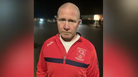 Paul is looking into the camera. He's wearing a red O'Neills football shirt with a Drumconrath logo. It's nighttime and he's outside. 