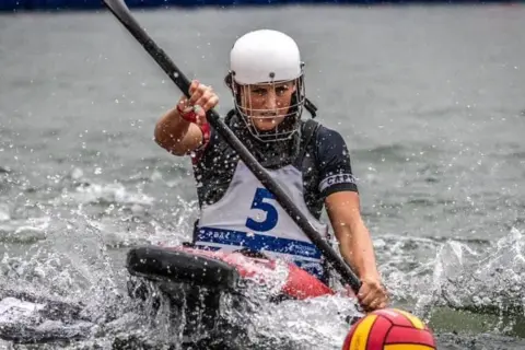 David McBay/Paddle UK A woman in a canoe on water. She is wearing a black top and a blue and white vest with the number 5 on it. Her canoe is red, the paddle she is using is black and there is a yellow and red ball in front of her which she is looking at with a face of concentration. She is also wearing a white helmet.