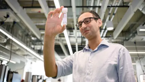 Cambridge University Hospitals/Mel Yeneralski Dr Tadross is in a lab. He has short dark hair, black framed glasses and is wearing a pale blue shirt with rolled up sleeves. In his right hand, he is holding up a transparent specimen tube containing clear fluid. 