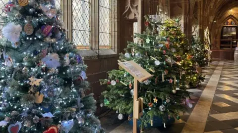 BBC A row of decorated Christmas trees in a cathedral corridor, with a brown-and- cream tiled floor and stone-framed windows.