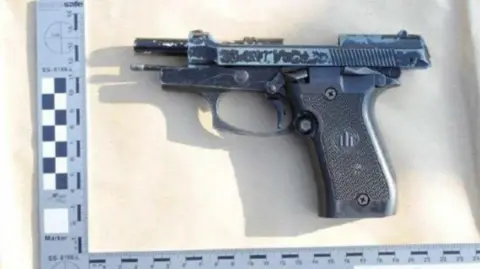 Staffordshire Police A gun, with a ruler below and above to measure its size. It is black, with metal visible as some of the paint has chipped off.