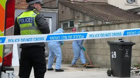 An officer stands behind police tape as  forensics in blue scrubs examine the scene . 