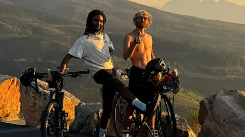 Contributed Two young men stand in front of a grassy hilly area and smile at the camera. One wears a white T-shirt and has black dreadlocks. He holds his bike in one hand while resting his other arm behind his back. The other has curly blonde hair, is shirtless, has glasses on and raises his thumb. He sits on top of his bike.
