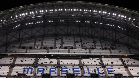Reuters Inside the Stade Vélodrome showing one of the stands. The stand has a curved roof and is filled with white chairs. Towards the bottom Marseille is spelt out in blue chairs. Groups of people are sat in their seats but it is largely empty. It is dark and the stadium lights are on.
