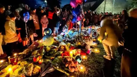 Scores of flowers and candles left by the crash. It is evening and there are loads of people standing around the flowers and candles that have have been left.