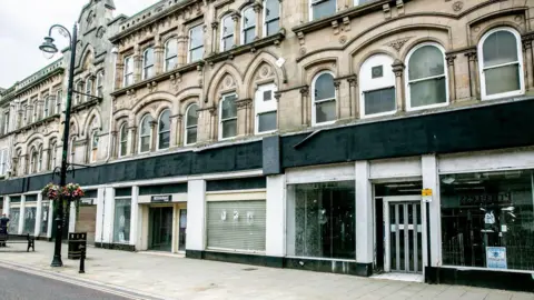 80 Newgate Street. The former department store stands vacant. Shutters are down over some of its windows and the former shop sign running along the length of the building has been painted black. The upper floors of the building are made of ornate stonework.