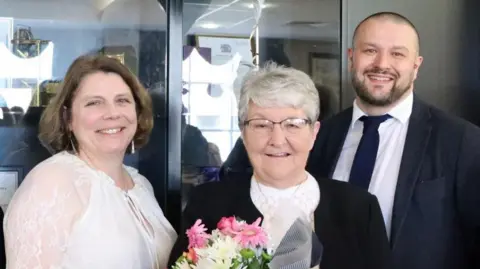 A group of two women and one man stand together for a picture. The woman in the centre is holding a bouquet of flowers.