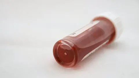 A vial of blood closed with a white plastic cap lies on its side on a white table 