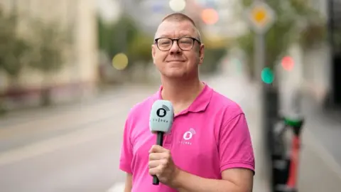 En mann i en rosa poloskjorte står på en gate og holder en mikrofon mot brystet. Han bruker briller og smiler stolt,